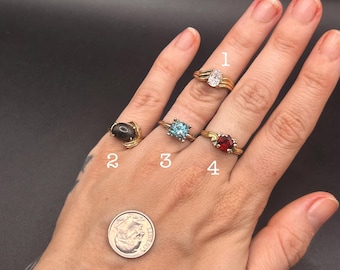 May include: A hand displays four gold rings with various gemstones: an oval white stone, a black stone, a blue stone, and a red stone. A dime is also present. The rings are numbered 1 through 4.