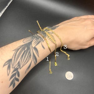 May include: Three gold-colored bracelets are displayed on a wrist with a floral tattoo. The bracelets include a delicate chain, a thin bar, and a chain with a decorative charm. A coin is also visible.