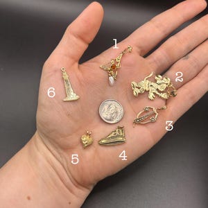 May include: Six gold-toned metal charms labeled with numbers one through six, including a lighthouse, a heart, a shoe, a cartoon mouse, and two decorative pendants with small stones, arranged around a United States dime for scale.