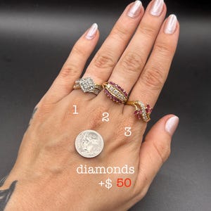 May include: Three gold rings labeled 1, 2, and 3. Ring 1 features a cluster of diamonds. Ring 2 has a row of diamonds flanked by red stones. Ring 3 displays a leaf-like design with red stones and diamonds. A US dime is shown for scale.