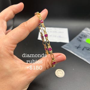 May include: A gold-toned bracelet featuring a series of oval-cut red rubies and small clear diamonds set in a geometric metal link pattern. The text "diamonds rubies +$150" is visible on the image, along with a small silver-colored coin.