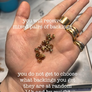 May include: A hand holding a collection of small, gold-colored earring backs. The text on the image states that the backs are 14k gold and are selected at random. Several rings are visible on the fingers.