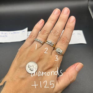 May include: Three gold rings with diamond accents worn on fingers. Ring one is a thin band with small diamonds. Ring two is a two-piece set with a central diamond. Ring three features a bypass design with two diamonds. A US dime is visible for scale.