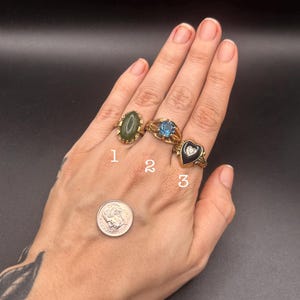 May include: Three gold rings with gemstones are displayed on a hand. Ring 1 has an oval green stone, ring 2 has a blue stone, and ring 3 has a black heart-shaped stone with a white center. A dime is also present.