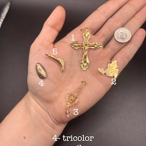 May include: A collection of gold-toned jewelry pieces, including a cross pendant, a map-shaped charm, a pendant with pearls and crystals, a curved piece, and a tricolor nail. The items are displayed on a dark background.