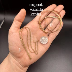 May include: A hand displays two gold necklaces, a set of gold bracelets, and a dime. The necklaces are delicate chain styles. The bracelets are circular, with one having a textured surface. The text "expect vanilla kinks" is visible.