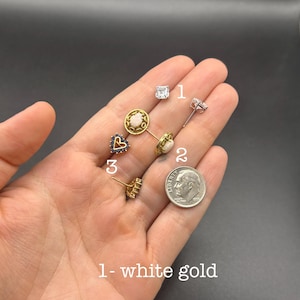 May include: Three distinct stud earrings are displayed. Number 1 is a square white gold earring with a clear stone. Number 2 features a round gold setting with a white stone. Number 3 is a heart-shaped gold earring with blue stones, next to a US dime.