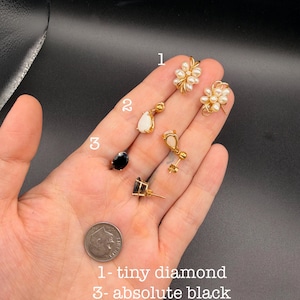 May include: Three pairs of gold earrings labeled 1, 2, and 3. Pair 1 features clusters of small white pearls. Pair 2 consists of teardrop-shaped white stones. Pair 3 features oval-shaped black stones. A US dime is included for scale.