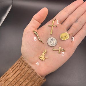 May include: Five gold-toned pendants labeled 1 through 5, including a cross, a circular religious medallion, a smaller cross, a cat figure, and a curved horn-shaped charm, arranged around a silver-colored United States dime for scale.