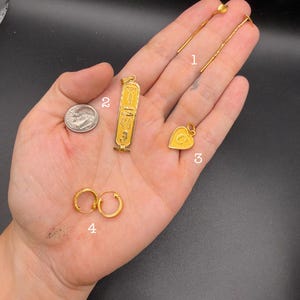May include: A collection of gold-toned jewelry items including a pair of long threader earrings, a rectangular pendant with Egyptian-style hieroglyphics, a heart-shaped pendant with the letter G, and a pair of small hoop earrings next to a US dime.