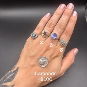 May include: Four gold rings featuring various gemstones including a cluster ring, a ruby ring with diamond accents, a large oval blue stone ring, and a heart-shaped pavé ring, displayed with a US dime for scale and the text diamonds +$100.
