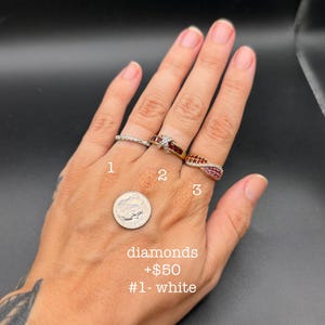 May include: Three rings on fingers: ring 1 is a thin silver-tone band with small clear stones, ring 2 is a gold-tone band with red stones and a silver-tone cross detail, and ring 3 is a twisted gold-tone band with pink and orange stones.