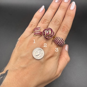 May include: Three gold rings featuring channel-set red stones, labeled 1, 2, and 3. Ring 1 has a curved design, ring 2 is a circular floral shape with a center stone, and ring 3 is an oval dome. A US dime is shown for scale next to the rings.