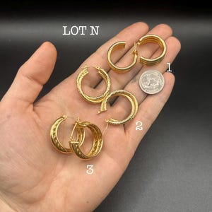 May include: A selection of gold-colored hoop earrings, each with a unique design. The earrings are displayed on a hand against a dark background. The text "LOT N" is visible in the upper left corner. Three pairs of earrings are numbered 1, 2, and 3.