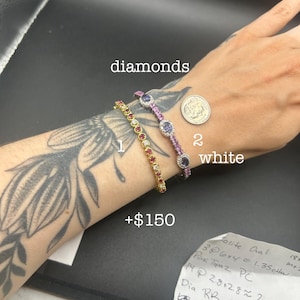 May include: Two bracelets on a wrist next to a US dime for scale. Bracelet 1 is a yellow gold chain with red stones and diamonds. Bracelet 2 is a white metal chain with oval purple stones surrounded by small diamonds. Text labels include diamonds, 2, white, and +$150.