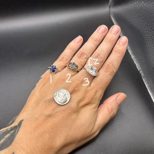 May include: Three rings worn on fingers, labeled 1, 2, and 3. Ring 1 has a blue stone, ring 2 has three dark stones, and ring 3 is a large clear stone labeled CZ. A US dime coin is placed on the hand for scale, showing the relative size of the rings.