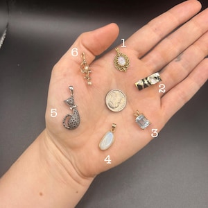 May include: Six distinct jewelry pendants labeled one through six, including an oval opal pendant, a rectangular black and gold bar, a rectangular blue gemstone, an oval moonstone, a cat-shaped pendant, and a pearl drop, alongside a US dime.