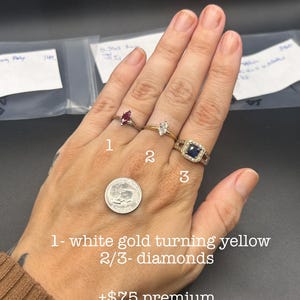 May include: Three rings worn on fingers: a marquise-cut red gemstone ring, a marquise-cut clear gemstone ring on a gold band, and a square-cut blue gemstone ring with a diamond halo. A US dime coin is placed on the hand for size reference.