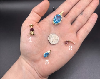 May include: A collection of jewelry pieces, including a gold pendant with a large oval blue gemstone, a small gold earring with a pink gemstone, and a teardrop-shaped blue gemstone pendant. A small gold candlestick with a red gemstone is also present.