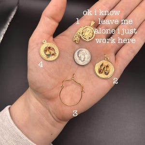 May include: A collection of gold-toned charms and a dime. The charms include a Native American profile, a pair of ruby slippers, a decorative open circle, and a charm with a cross and green stone. Text on the image reads: "ok i know leave me alone i just work here."