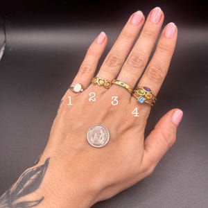 May include: Four gold rings with gemstones and a coin are displayed on a hand against a black background. The rings feature various designs, including a ring with a white stone and a ring with multiple colored gemstones.
