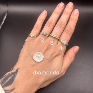 May include: Four gold rings with diamond accents are displayed on a hand. The rings include a knot design, a starburst, a band with small stones, and a round cluster. A dime and the word "diamonds" are also visible.