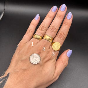 May include: A hand displaying three gold rings and a silver coin. Ring 1 is a simple band, ring 2 has a textured design, and ring 3 is a signet ring. The coin is a dime. The background is black.