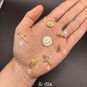 May include: A collection of gold-colored charms, including crosses, a circular pendant, a letter "A" charm, and a charm with three small stones. The charms are displayed on a dark background. The text "2-dia" is visible.