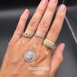 May include: Three rings on fingers: ring 1 is a gold band with two rows of small red stones and clear stones, ring 2 is a thin silver band with clear stones, and ring 3 is a wide gold band with five clear stones. A US dime is shown for scale.