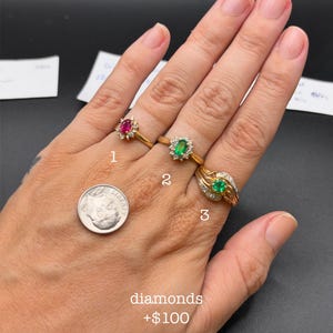May include: Three gold rings worn on fingers. Ring 1 features a central oval ruby surrounded by small diamonds. Ring 2 has a central oval emerald with a diamond halo. Ring 3 displays a round emerald flanked by diamond-accented gold ribbons.