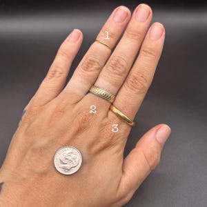 May include: Three gold-toned rings labeled 1, 2, and 3. Ring 1 is a thin band, ring 2 features a textured braided pattern, and ring 3 is a smooth, thick band. A silver-colored US dime coin is positioned near the rings for scale reference.