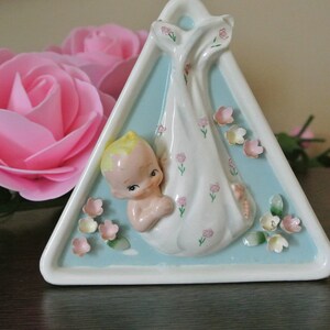May include: A white ceramic triangular trinket dish featuring a baby doll figurine. The doll is dressed in a white dress with pink flowers and holds a pink flower. The dish is decorated with pink flowers and has a light blue background.