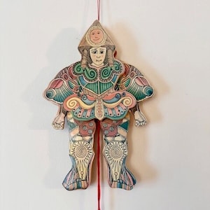 May include: A colorful wooden puppet with a whimsical design. The puppet is dressed in a multi-colored outfit with intricate patterns and a pointed hat. The puppet is suspended from a red string with a red ball at the end.