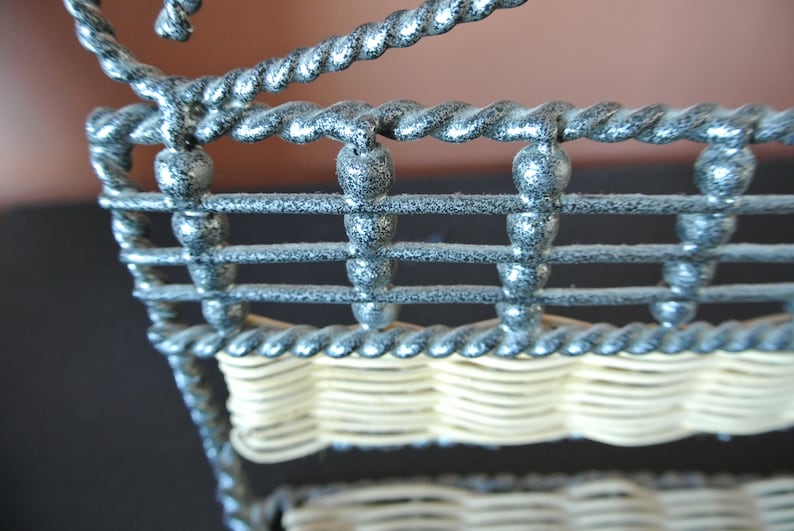 Pu&ograve; includere: Primo piano di una rastrelliera in filo metallico argentato con un fondo in vimini intrecciato. La rastrelliera ha un design a corda decorativo ed &egrave; realizzata in filo ritorto.