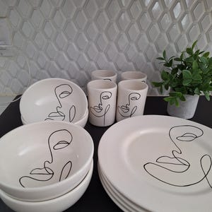 May include: A collection of white ceramic tableware with a minimalist black line art face design. The set includes bowls, cups, and plates. A small potted plant is visible in the background.