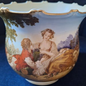 Peut inclure: Un pot en porcelaine blanche avec un bord doré et une scène peinte de deux personnes dans un jardin. Le pot a un bord festonné et une base ronde.