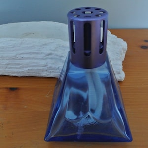 May include: A blue glass diffuser bottle with a purple metal top and a wick. The bottle is shaped like a pyramid and has a decorative design on the front.