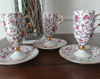 Könnte beinhalten: Set aus drei weißen Porzellan-Teetassen und Untertassen mit einem floralen Muster in Rosa-, Grün- und Goldtönen. Die Tassen haben einen goldenen Rand und einen goldenen Henkel.