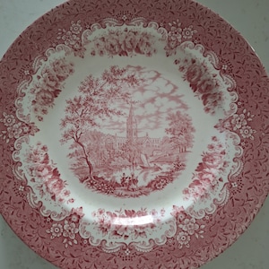 Peut inclure: Une assiette en céramique rose et blanche avec une bordure florale et une image centrale d'un bâtiment avec des arbres et une rivière. L'assiette est décorée d'un motif floral délicat.