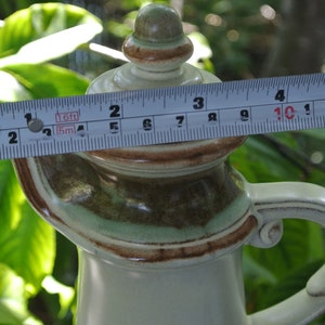 Pode incluir: Uma jarra de cer&acirc;mica com um esmalte castanho, verde e branco. A jarra tem uma tampa com um bot&atilde;o no topo. A jarra tem 25,4 cm de altura.
