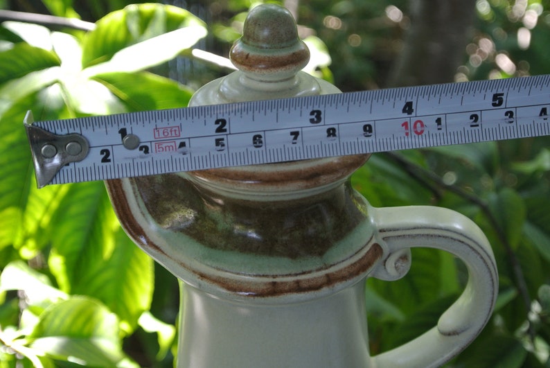 Pode incluir: Uma jarra de cer&acirc;mica com um esmalte castanho, verde e branco. A jarra tem uma tampa com um bot&atilde;o no topo. A jarra tem 25,4 cm de altura.