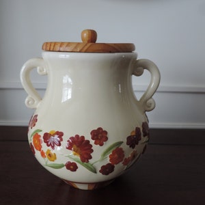 May include: A white ceramic cookie jar with a wooden lid. The jar is decorated with a floral pattern in shades of red, orange, and yellow.