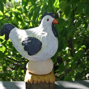 Può includere: Una statuetta di pollo in ceramica bianca e nera in piedi su un uovo grande con una base di paglia. Il pollo ha un becco rosso e occhi neri.