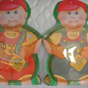 Puede incluir: Dos conjuntos de muñecas Cabbage Patch Kids en un embalaje de cartón. Los conjuntos son un conjunto de bombero rojo y amarillo y un conjunto de mono vaquero azul. Ambos conjuntos tienen una camiseta amarilla debajo. El embalaje tiene el logotipo de Cabbage Patch Kids y las palabras "Romper" y "Cabbage Patch Kids".