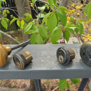 Può includere: Quattro pomelli e serrature per porte antiche, due con pomelli in legno, uno con pomo in ottone e uno con pomo in metallo nero. I pomelli sono su una superficie in legno grigia.