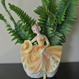 May include: A ceramic planter shaped like a woman in a yellow and orange dress holding a hat and flowers. The planter is filled with green ferns.