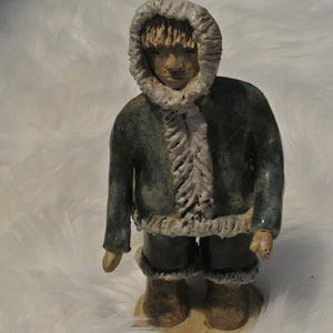 Puede incluir: Una figurita de cerámica de una persona que lleva una chaqueta azul con capucha y ribete de piel blanca. La figurita está de pie sobre una superficie blanca.