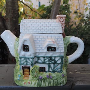 Può includere: Una teiera in ceramica bianca a forma di cottage con finiture verdi, una porta marrone e un tetto bianco. La teiera ha un manico verde e marrone e un beccuccio bianco.