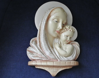 May include: A white ceramic wall sculpture of the Virgin Mary holding the baby Jesus. The sculpture is in a relief style and has a gold-coloured halo around Mary's head.