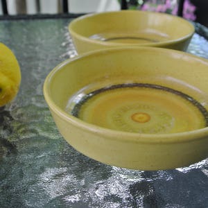 Puede incluir: Dos cuencos de cer&aacute;mica amarillos con un borde marr&oacute;n y un dise&ntilde;o de remolino amarillo y naranja en el fondo. Los cuencos est&aacute;n sobre una superficie gris texturizada.